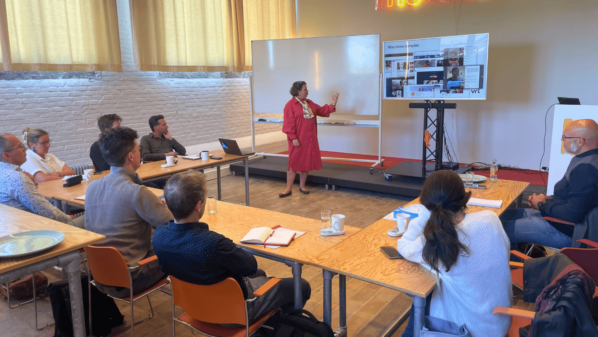Presenter in red dress points to whiteboard during a professional meeting with attentive audience.