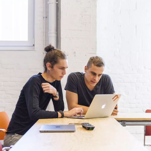 Two men collaborate on a laptop at a light wood table in a modern office.