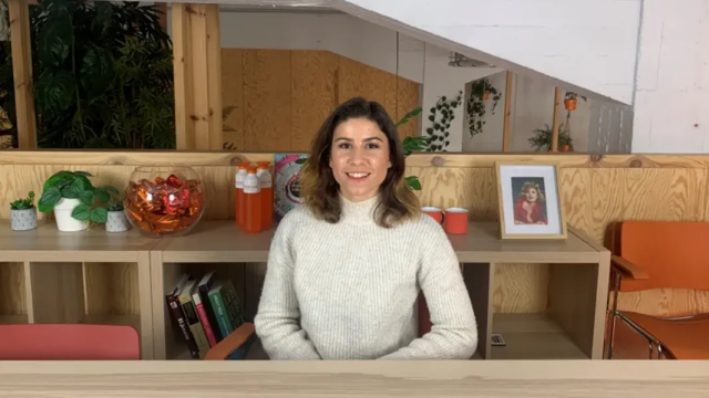 Woman in a white sweater smiling at a desk with plants and orange accessories.