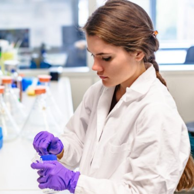 Scientist in lab coat and purple gloves opening a chemical container in a research lab.